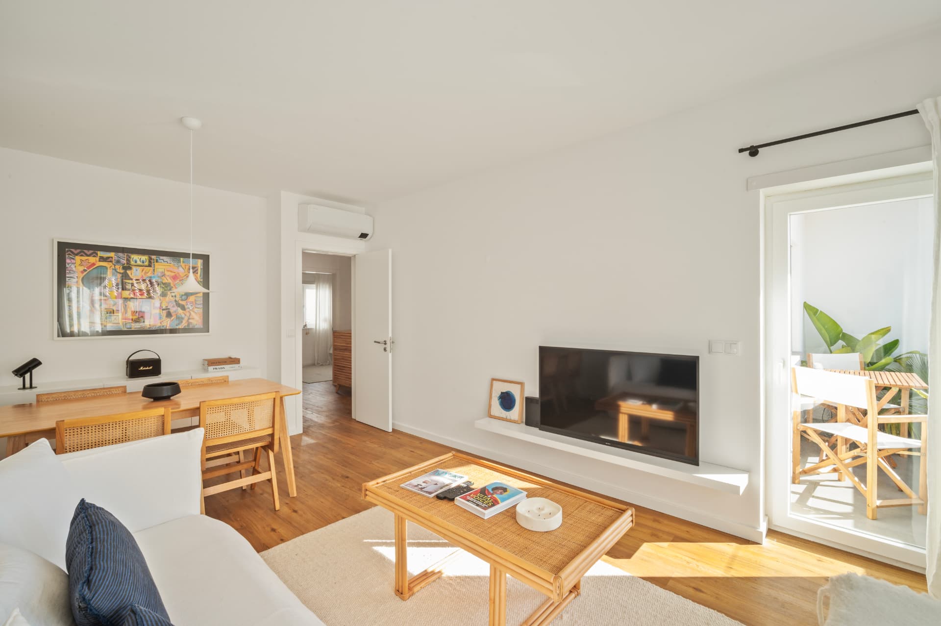 The living room: a white sofa and a low coffee table on a pale rug, the dining table and cane chairs to the left, and the sunlit terrace door to the right