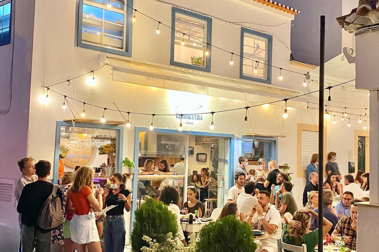 A restaurant terrace in Cascais at night, festoon lights overhead and guests dining at tables below the lit sign